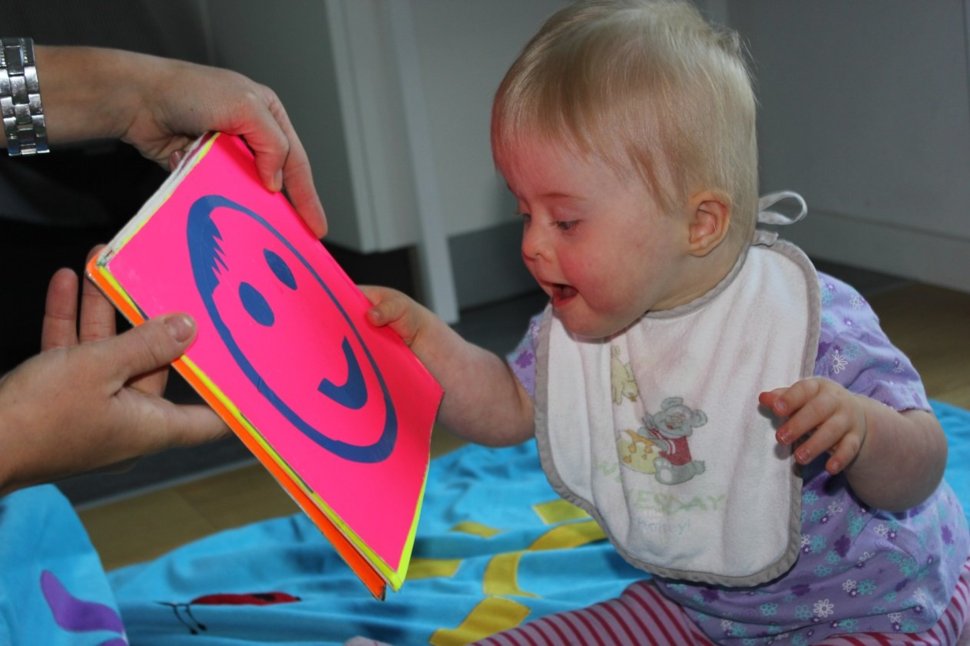 Ein lächelndes Kleinkind mit einem Lätzchen betrachtet freudig ein buntes Buch mit einem großen Smiley-Gesicht.