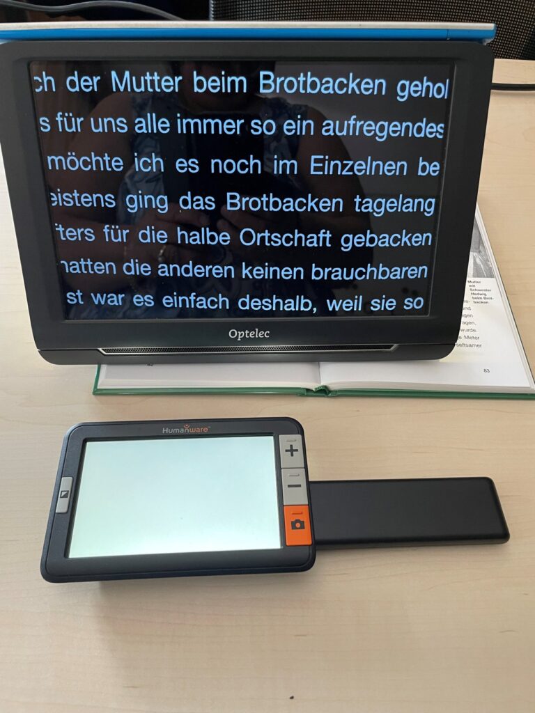 Ein elektronisches Vergrößerungsgerät zeigt eine vergrößerte Textseite an, unterstützt durch ein tragbares Lesegerät daneben, zur Unterstützung von sehbehinderten Personen.