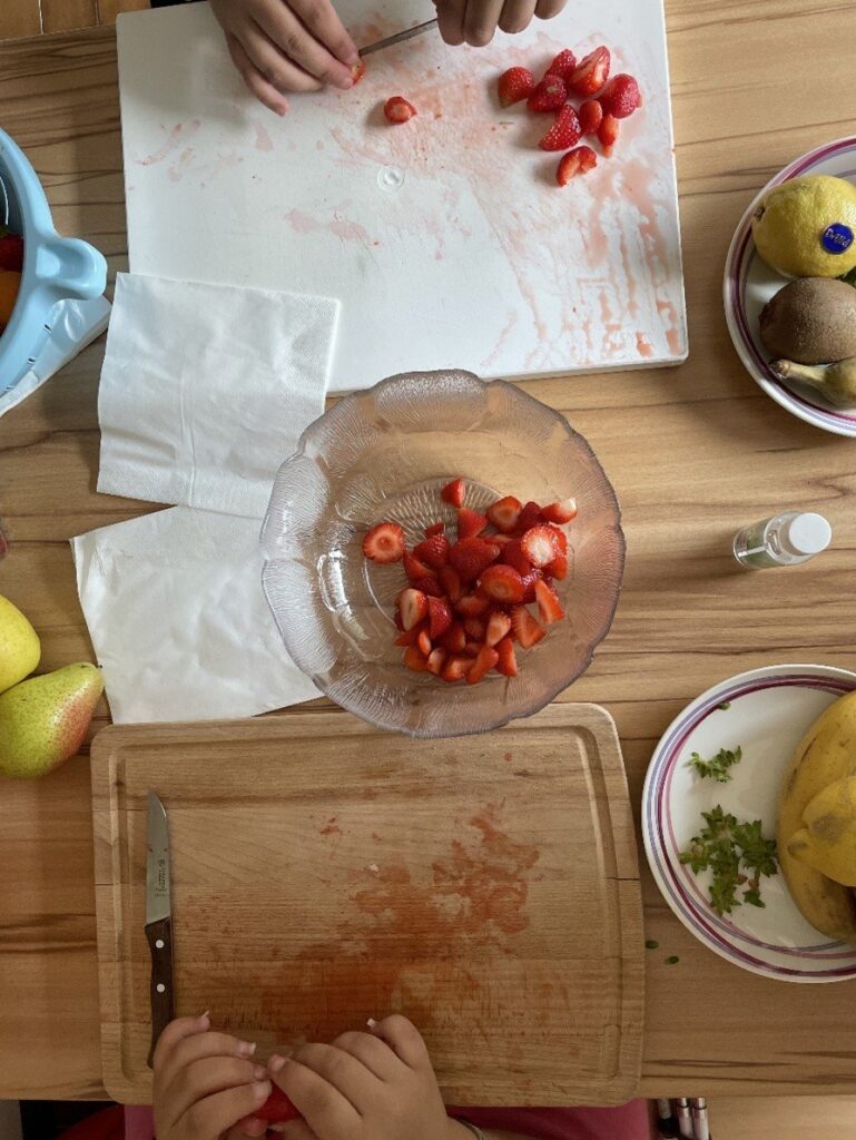 Zwei Personen schneiden Erdbeeren auf Schneidebrettern, umgeben von Obst und Küchenutensilien auf einem Holztisch.