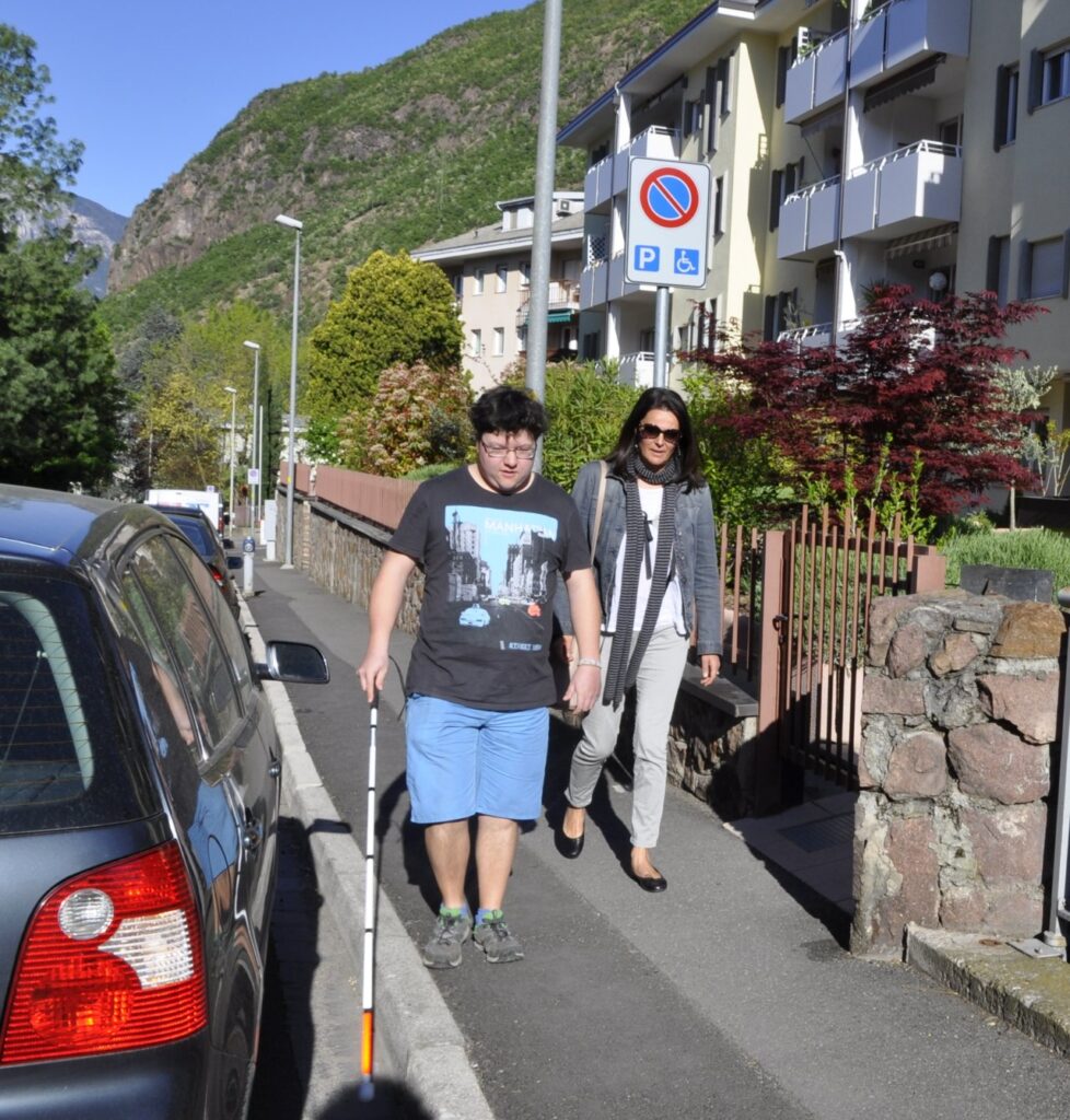Ein Mann mit einem Blindenstock geht gemeinsam mit einer Frau auf einem Bürgersteig entlang, der durch ein Schild als barrierefrei gekennzeichnet ist.
