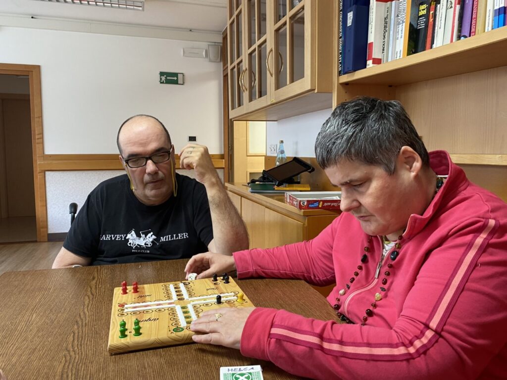 Zwei Personen spielen gemeinsam ein Brettspiel an einem Holztisch in einem Raum mit Bücherregalen.