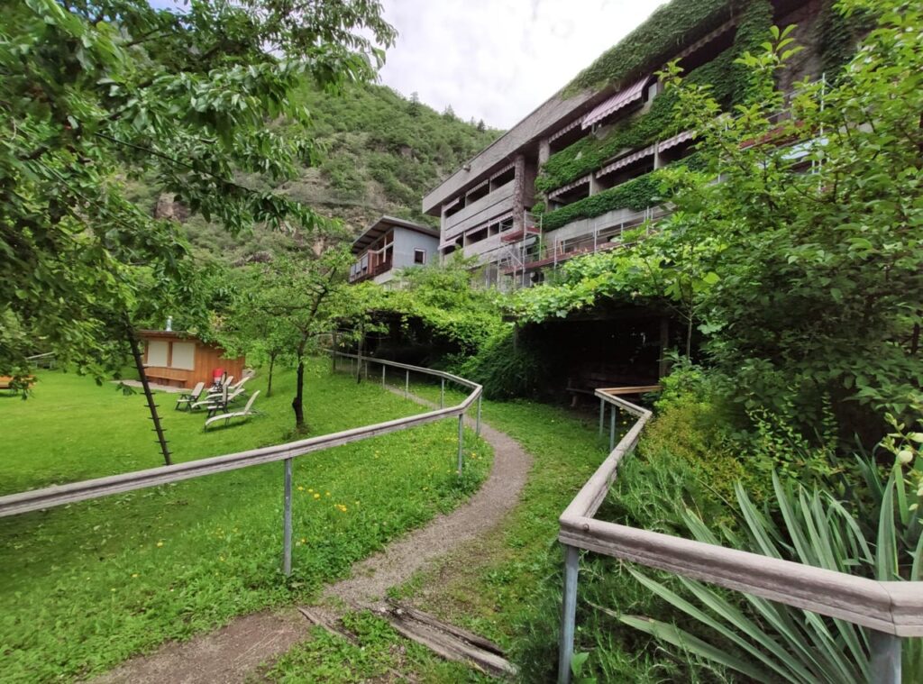 Eine gewundene, leicht begehbare Rampe mit Handlauf führt zu einem von Pflanzen umgebenen Gebäude.