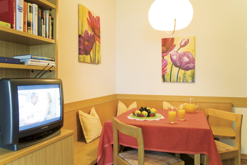 Ein kleiner Essbereich mit rotem Tischtuch, Obst, Getränken und einem eingeschalteten Fernseher in einem gemütlichen Raum mit Blumenbildern an der Wand.