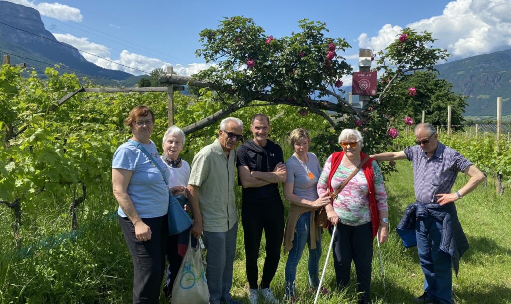 Eine Gruppe von Menschen steht lächelnd in einem sonnigen Weinberg vor einem blühenden Baum, mit Bergen im Hintergrund.