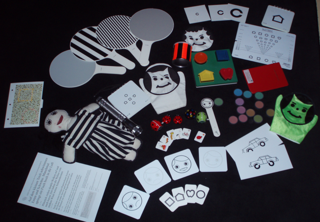 Das Bild zeigt eine Sammlung von Förderspielzeug und Karten für visuelle oder kognitive Tests, darunter Handpuppen, farbige Chips, Musterkarten und Plüschfiguren auf einem dunklen Hintergrund.