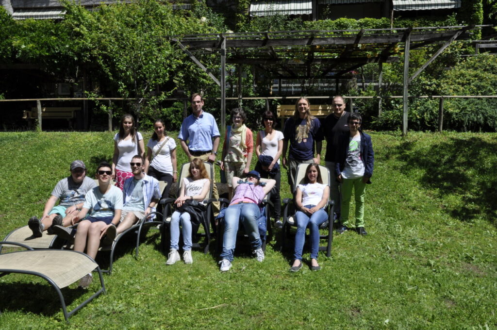 Eine Gruppe von Menschen sitzt und steht auf einer grünen Wiese vor einer natürlichen Kulisse.