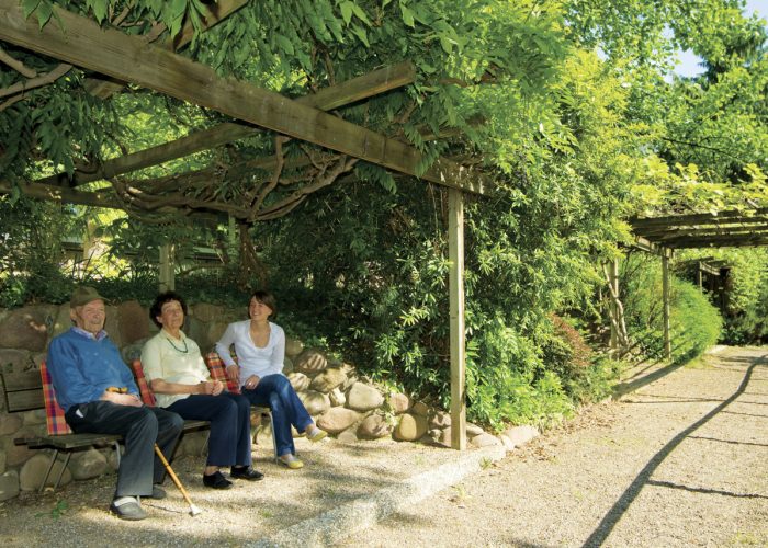 Drei Personen sitzen unter einer schattigen Laube auf einer Holzbank in einem grünen, sonnigen Garten.